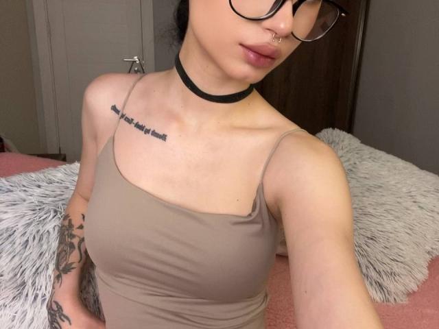 GlitchDoll con gafas, aro en la nariz, collar negro, camiseta marrón, tatuaje en el pecho, tatuaje en el brazo, sentada en una cama rosa y blanca. Se ven los senos, los pechos son pequeños..