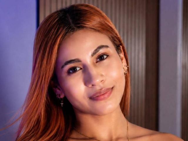 Ariana Wright tem a pele clara, cabelos lisos e alaranjados, pequenos brincos e um sorriso discreto. Ela está usando um colar fino de ouro, e a iluminação em seu rosto é suave e quente..