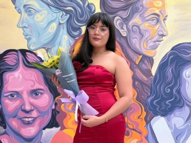 EleanorB, a chubby woman with black hair and glasses, wears a strapless red dress, holding a bouquet of yellow flowers, standing in front of colorful painted portraits.