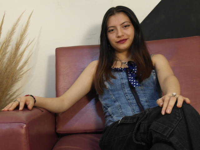 Samantha Ferrero, con cabello largo y castaño, vestida con una blusa azul de mezclilla sin mangas y pantalones negros, sentada en un sofá de cuero rojo, sonriendo, con los brazos apoyados en el sofá..
