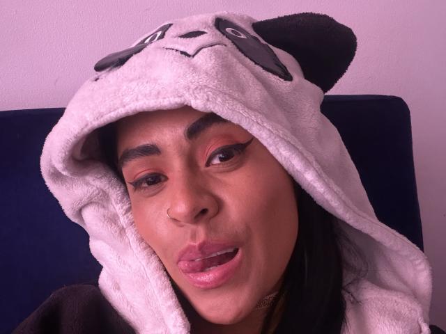 StellaBeckker with dark skin, pink lipstick, and a cute panda hat, sticking her tongue out, wearing red eyeliner, and a nose ring. Soft pink lighting.
