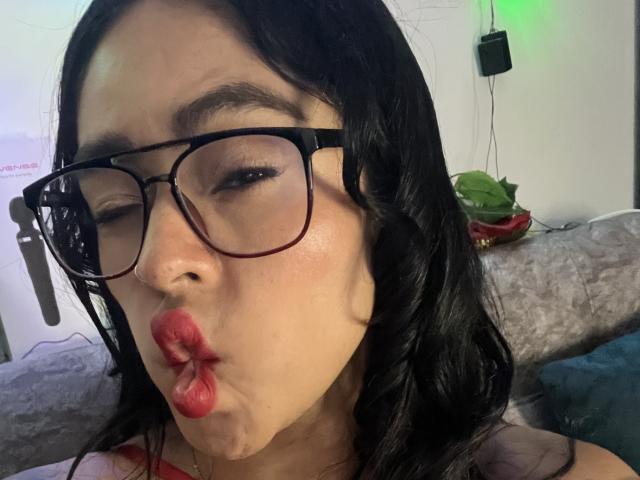 StellaBeckker, con i capelli neri e ondulati, rossetto rosso e occhiali, che fa un'espressione civettuola. Le sue natiche non sono visibili. Ha una carnagione chiara..