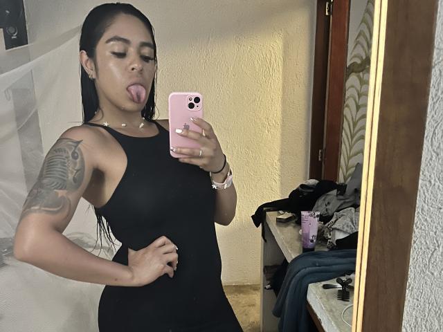 StellaBeckker in a black dress, tongue out, holding a pink phone, tattoo on right arm, mirror selfie, medium tits, hand on hip, wet black hair, bathroom.