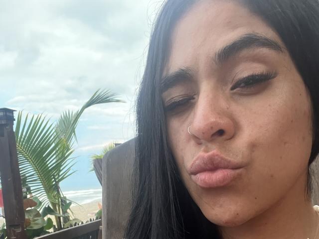 StellaBeckker, con cabello negro, un piercing en la nariz y labios rosados y fruncidos, cejas marrones y pobladas, y pestañas largas, mostrando una ligera sonrisa. En el fondo, se ve una playa y una palmera..