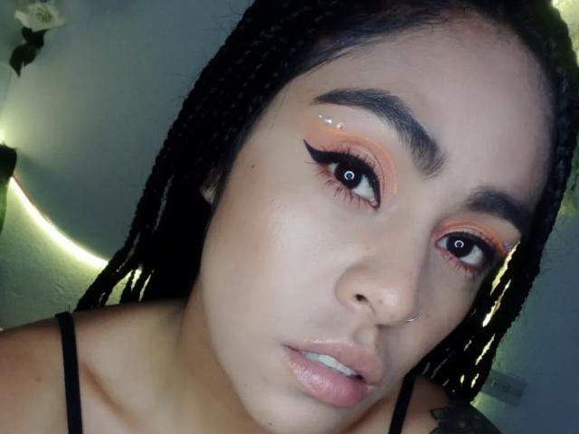 Il viso di StellaBeckker, con ombretto arancione lucido, eyeliner nero marcato e labbra rosa piene. Ha i capelli neri intrecciati, un anello al naso e un tatuaggio discreto sulla spalla..
