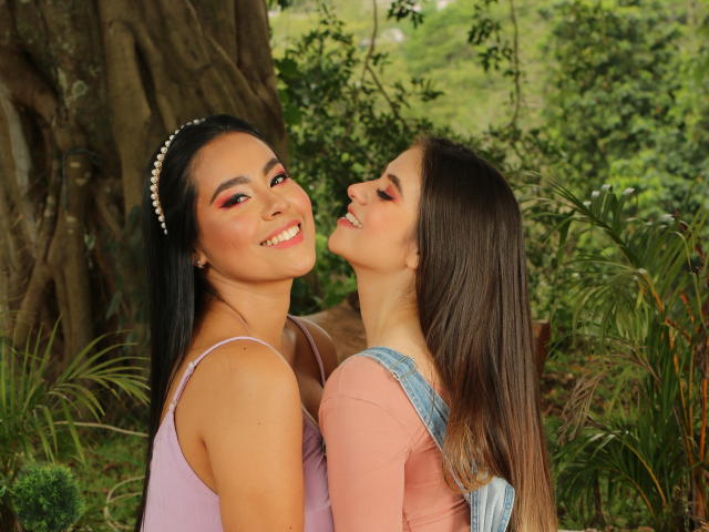 PaolaYKatty con i capelli neri e un vestito rosa, sorride a PaolaYKatty con i capelli castani, che indossa una maglietta rosa, ed è china in avanti, all'aperto vicino a un grande albero; entrambe mostrano espressioni carine e felici..