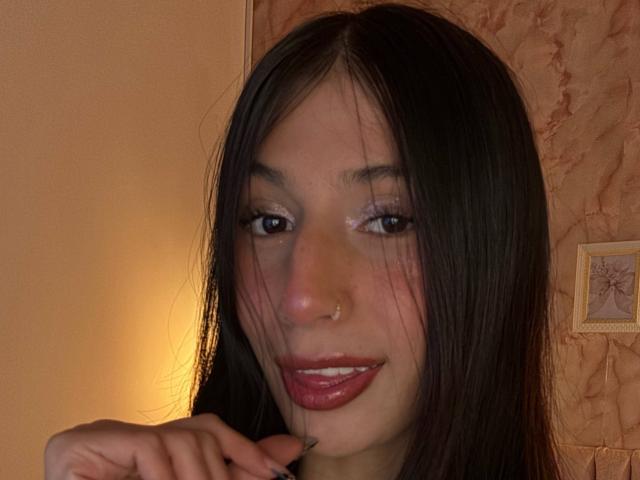 Sophie Beaumont tem cabelo preto liso, um pequeno piercing no nariz, lábios rosados brilhantes e olhos castanhos, com um sorriso discreto e uma mão perto do queixo..
