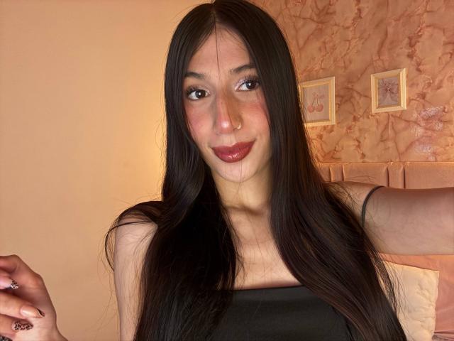 Sophie Beaumont a de longs cheveux noirs, un petit anneau au nez et un sourire discret. Elle porte un haut noir qui dévoile sa poitrine, avec du rouge à lèvres rose et un maquillage léger..