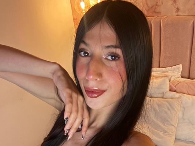 Sophie Beaumont, com longos cabelos pretos, lábios rosados e brilhantes, um piercing no nariz e um sorriso adorável, segurando o queixo com os dedos bem cuidados, em um quarto iluminado por uma luz quente. Suas nádegas não estão visíveis..