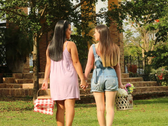 PaolaYKatty, con lunghi capelli neri e un vestito rosa, tiene la mano a PaolaYKatty, che indossa pantaloncini di jeans blu, entrambe portano cesti e sono in piedi sull'erba, vicino ad alberi e gradini in mattoni..