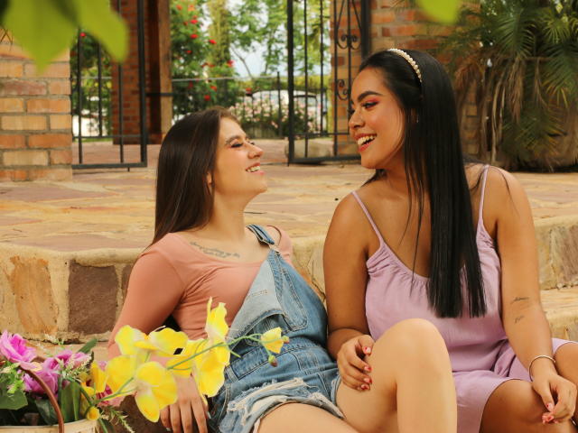 PaolaYKatty con lunghi capelli neri, sorridente, indossa una maglietta rosa e una tuta in denim, seduta accanto a PaolaYKatty che indossa un abito viola, tenendosi per mano, fiori gialli in primo piano..