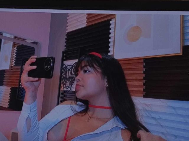 KimDevinne with long black hair, red choker, and red bra, taking a selfie in a mirror, wearing a blue shirt, showing her tits, in a dimly lit room.