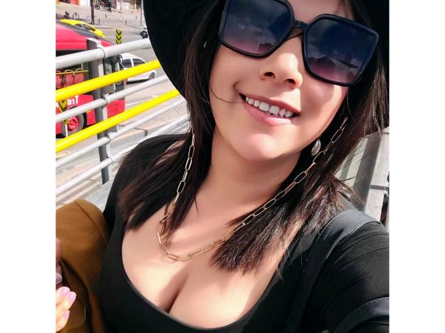 ZoeyGrey with dark hair, black sunglasses, and a hat, smiling, showing her cleavage in a black top, wearing a chain necklace, and holding a brown bag.