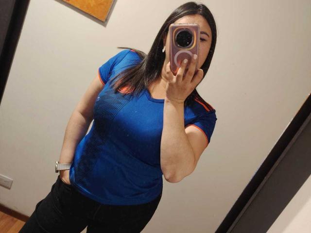 ZoeyGrey in a blue, tight-fitting t-shirt with red trim, black pants, and a silver watch, taking a mirror selfie with a purple phone, showing her tits and ass.