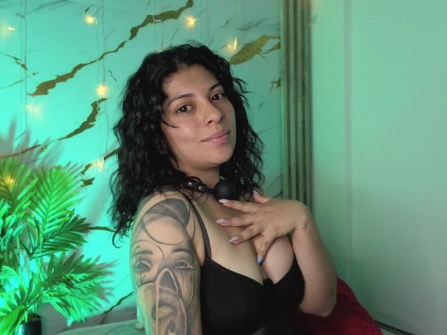 IsabellaFleur with curly black hair, wearing a black bra, showing off her tattooed arm and touching her chest. She has medium tits and a cute smile.