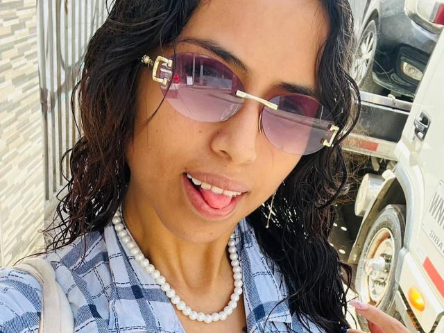 MyaaTyler with dark wavy hair, wearing pink sunglasses, a pearl necklace, and a blue plaid shirt, sticking her tongue out, showing her small, cute, pearly teeth.