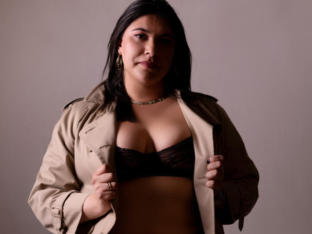 ZoeyGrey with tan jacket open, exposing black lace bra, holding jacket edges, wearing gold necklace, looking serious, dark hair, brown skin, medium tits, soft light, studio setting.