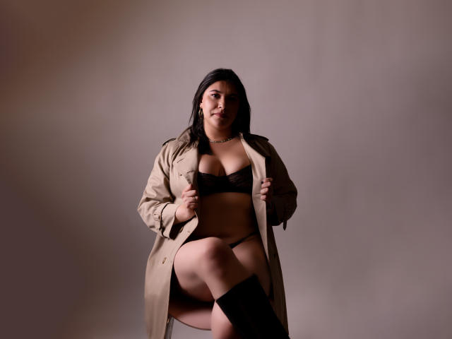 ZoeyGrey, curvy with dark hair, wearing a tan coat open to reveal black bra and panties, black knee socks, sitting with one leg up, smiling softly.