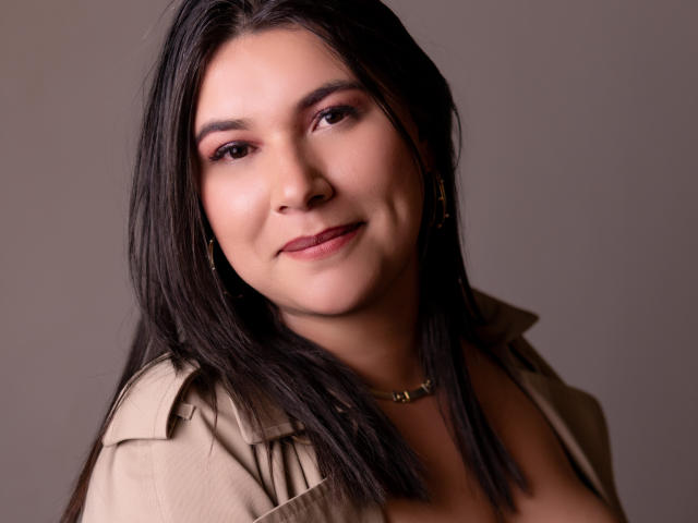ZoeyGrey has dark brown hair, pink eyeshadow, and a subtle smile. She's wearing a tan shirt with a necklace, and her small tits are slightly visible.