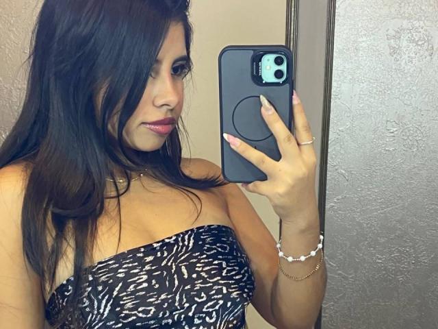 Celeste Morel, con i capelli scuri, il rossetto rosa e una canottiera nera e bianca senza spalline, tiene in mano un telefono nero in un selfie allo specchio, mostrando il suo seno e un anello..