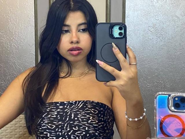 Celeste Morel, con lunghi capelli neri, rossetto rosa, indossa una canottiera nera e bianca a righe, sta scattando un selfie allo specchio con un iPhone blu, mostrando leggermente il seno..