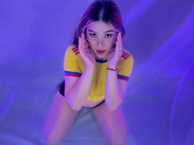 Elodie Gardner com um brilho roxo, camisa amarela com listras vermelhas, mãos nas bochechas, óculos, boca aberta, calcinha preta, ajoelhada, olhando para cima, cabelo castanho longo, pulseira vermelha, expressão de surpresa..