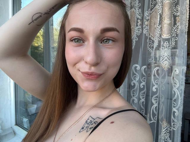 Cherelle Delozier mit langen braunen Haaren, grünen Augen und einem kleinen Nasenring, lächelnd, Hand im Haar, schwarzes Oberteil mit Trägern, Tattoo an der rechten Schulter und einer Halskette..
