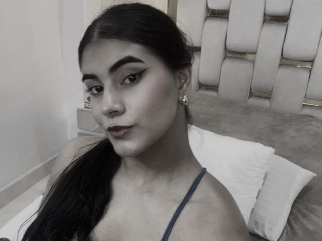 AlaraKey has long black hair, dark eyebrows, and wears dark lipstick. She's in a blue top, showing her tits, with a calm expression and small earrings.