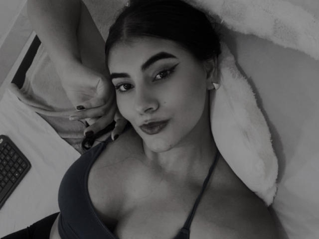 Black-and-white selfie of AlaraKey with dark hair, thick eyebrows, winged eyeliner, dark lipstick, and a tank top showing her big tits. Her hand rests above her head.