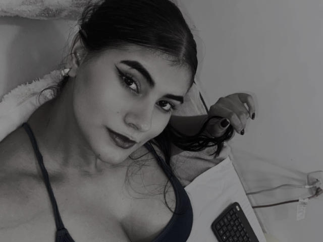 AlaraKey with dark hair, thick eyebrows, and dark lipstick, holding hair, wearing a low-cut top showing her tits, lying in bed with a phone and pillow. Black and white.