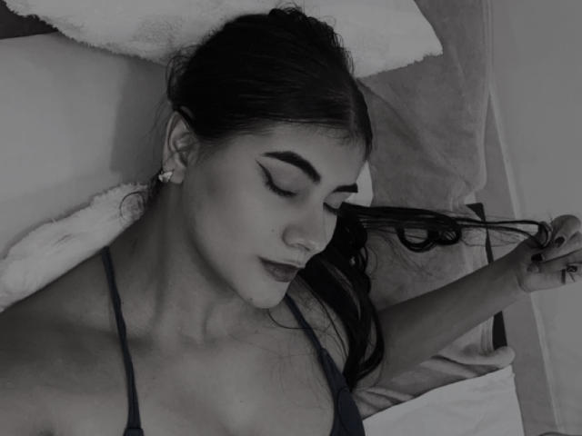 AlaraKey with dark hair, thick eyebrows, and dark lipstick, lying down, holding her hair, wearing a dark strap top, showing her small tits and a nose ring.
