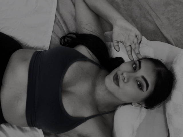 Black-and-white photo of AlaraKey lying on a bed, wearing a dark sports bra, showing her tits, with a nose ring, dark eyebrows, and long black hair, looking at camera.