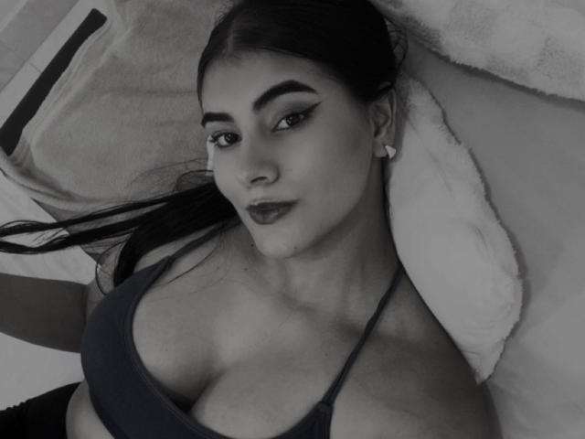 Black-and-white selfie of AlaraKey with dark lipstick, arched eyebrows, and small earrings, lying on a pillow, wearing a dark tank top, showing her boobs.