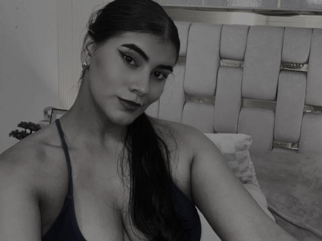Grayscale selfie of AlaraKey with dark hair in a ponytail, wearing a black halter top, showing her boobs, and dark lipstick, sitting on a bed with a tufted headboard.