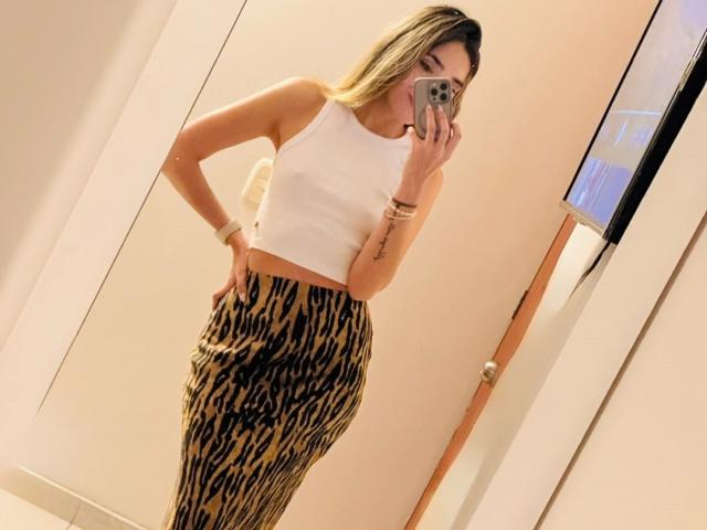 AdharaCooperr, avec ses cheveux blonds, portant un crop top blanc et un pantalon à imprimé léopard, tenant un téléphone, montrant une silhouette mince et des courbes, ainsi qu'un tatouage sur son bras droit..