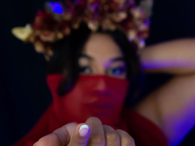 ValeryRiven with dark hair, red face mask, and flower crown, holding her finger with white nail polish, blue eye visible, blurry background, dark and blue lighting.