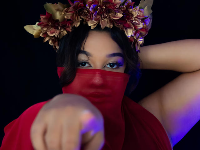 ValeryRiven with dark hair, wearing a red mask and headpiece, points forward with her finger, showing blue eye makeup against a dark background. She's dressed in a red top.