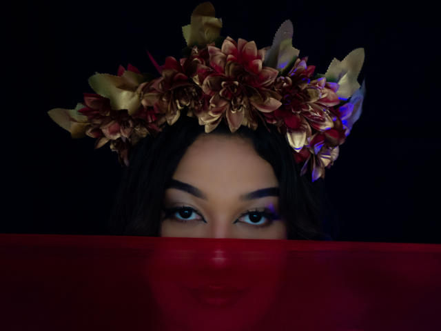 Das Gesicht von ValeryRiven mit dunklen Augen, sie trägt eine rote, durchsichtige Maske und einen Blumenkranz mit goldenen und roten Blütenblättern, vor einem dunklen Hintergrund. ValeryRivens schwarzes Haar ist sichtbar..
