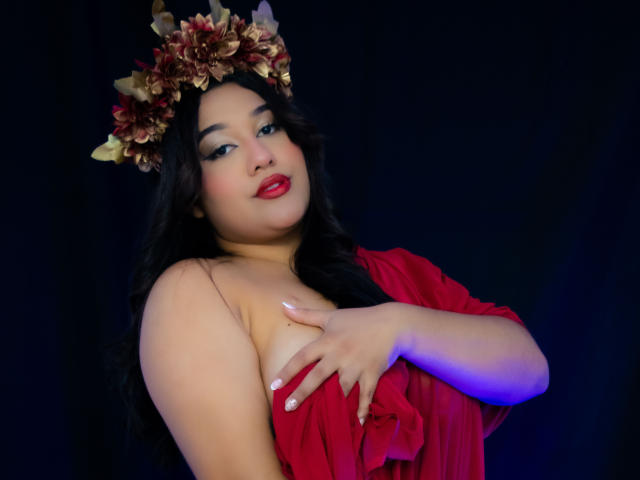 ValeryRiven with dark wavy hair, red dress, and flower crown, covering her tits with one hand, smiling with red lipstick, against a dark background, illuminated by blue light.
