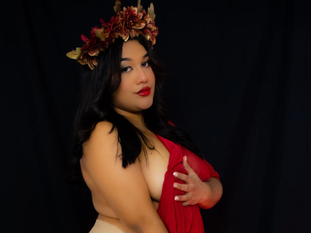 ValeryRiven with dark wavy hair, red lips, and a flower crown, wearing a red sheer dress, covering her big, round tits with one hand, against a black background.