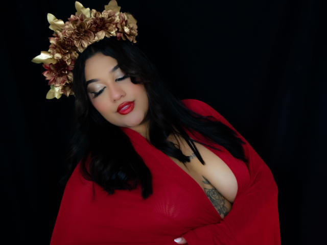 ValeryRiven with black hair, red dress, and gold flower crown, eyes closed, red lipstick, large boobs, tattoo on chest, arms crossed, dark background.