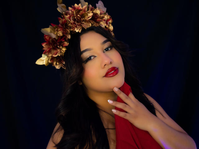 ValeryRiven, con cabello oscuro y ondulado, lápiz labial rojo, una corona de flores doradas y un vestido rojo, tocándose el cuello, fondo negro, iluminación suave, manos cerca del rostro, segura de sí misma, sexy..