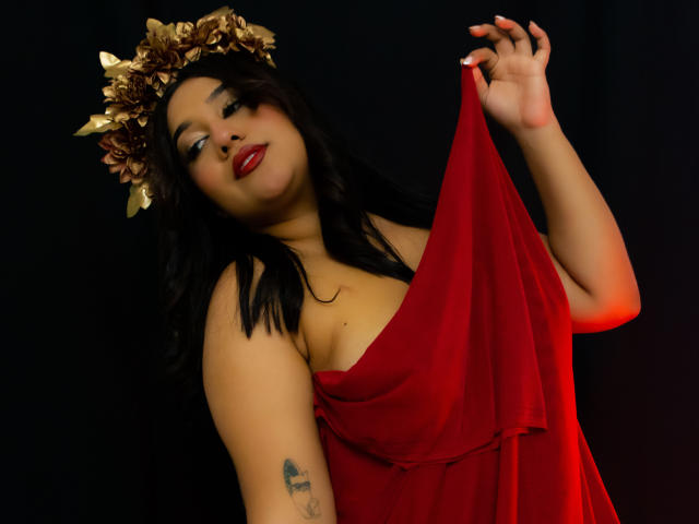 ValeryRiven, een Latina vrouw met lang zwart haar, gekleed in een rode jurk en een gouden bloemenkrans, houdt de jurk omhoog, toont een tatoeage op haar rechterarm, draagt rode lippenstift, op een donkere achtergrond..