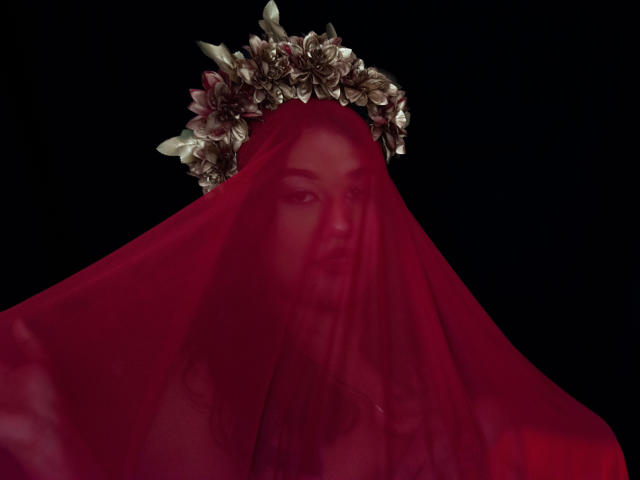 ValeryRiven met een rode sluier die haar gezicht bedekt, met een bloemenkrans om haar hoofd, zwarte achtergrond, de rode sluier onthult gedeeltelijk haar gezicht, de sluier is doorschijnend, de bloemenkrans heeft roze en witte bloemen..