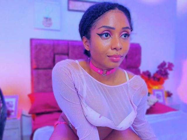 MeganWilliams with dark curly hair, pink choker, sheer white top, showing her small tits, sitting on bed, pink and purple lighting, looking at camera, slight smile, cute.