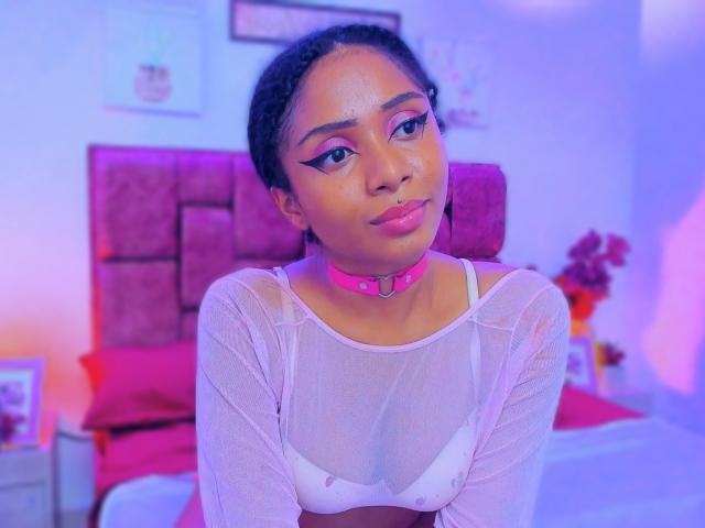 MeganWlliams with brown skin, pink choker, white sheer top, showing small white bra-covered tits, pink lipstick, and bold eyeliner, in a purple-lit bedroom.