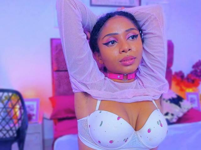 MeganWilliams with arms raised, wearing a pink collar, sheer pink top, and white bra with cherry patterns, exposing her big tits. She has dark skin, pink lips, and eyeliner.