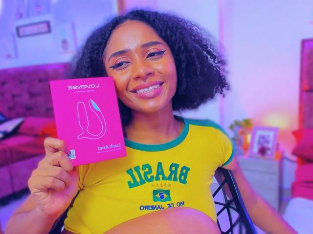 MeganWlliams with curly black hair, smiling, holding a pink card, wearing a yellow green Brazilian football shirt, showing a pregnant belly, in a brightly lit room.