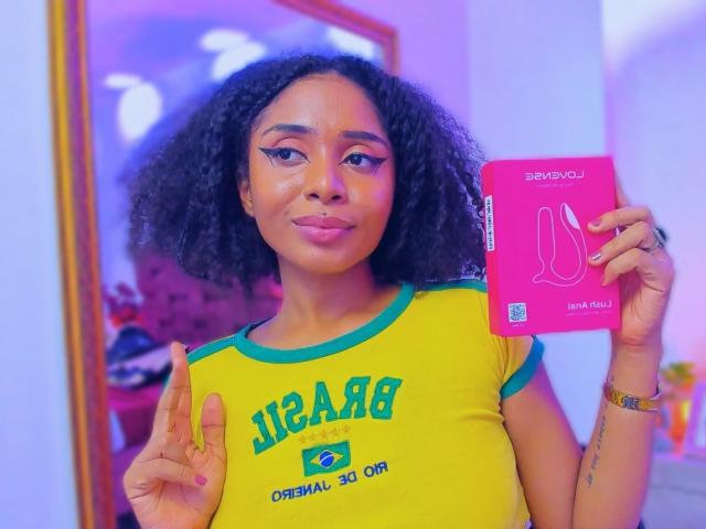 MeganWlliams with curly black hair, green and yellow shirt, holding pink sex toy box, making "rock on" hand gesture, pink and blue lighting, confident expression, pink lips.