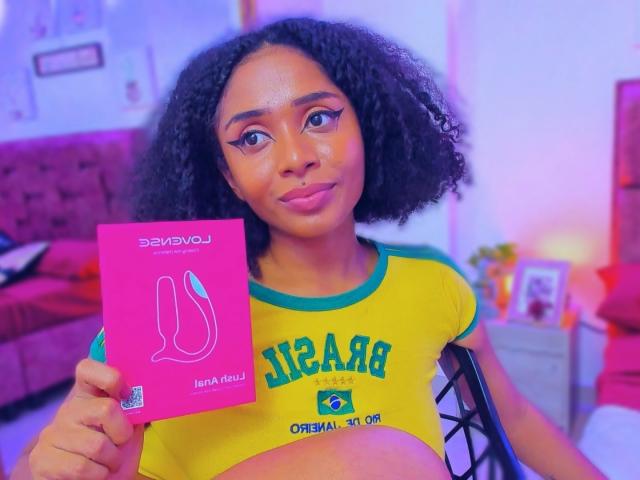 MeganWilliams with curly black hair, holding a pink sex toy box, wearing a yellow and green Brazilian-themed crop top, showcasing her tits. Her makeup is bold with winged eyeliner.